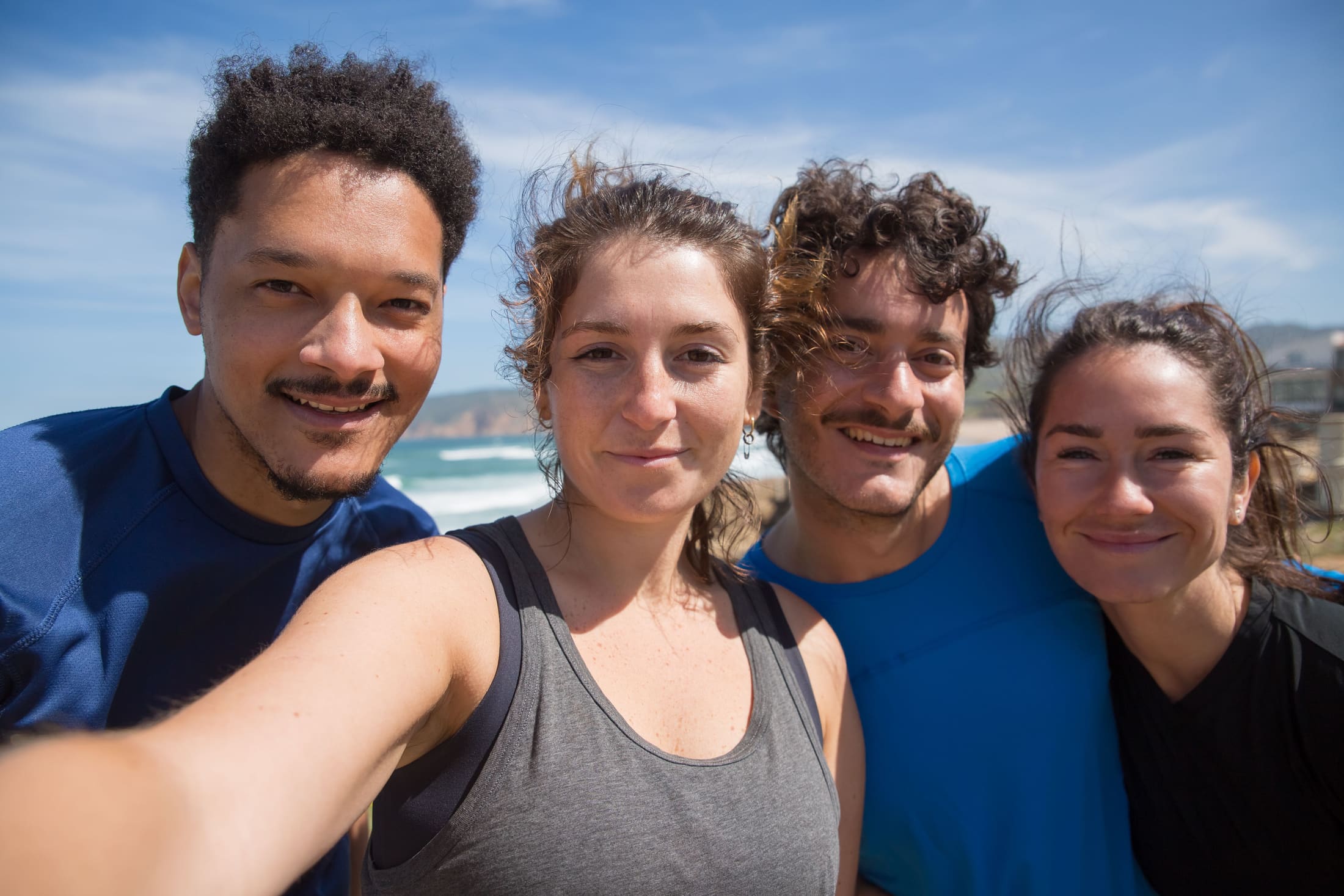 Four colleagues standing together outdoors on the coast in bright daylight, smiling at the camera