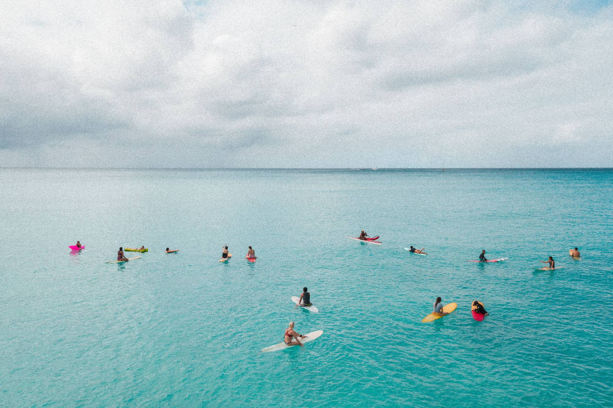 People on surfboards spread across calm turquoise ocean water, seen from above